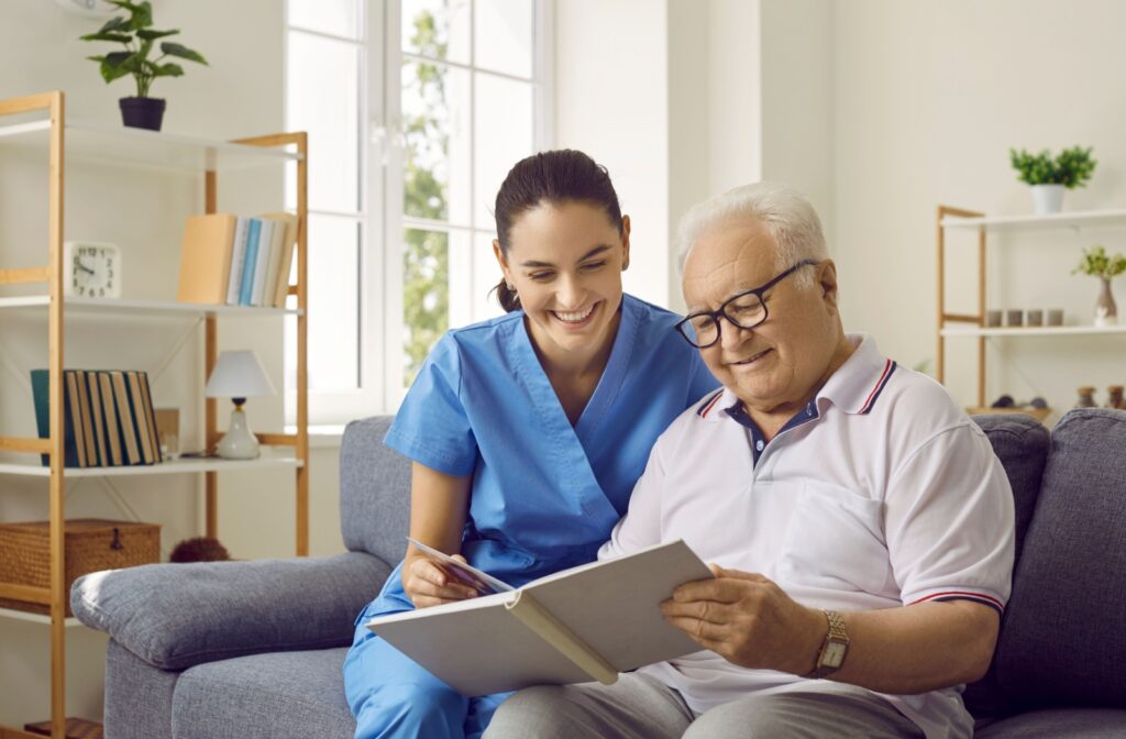 A memory care caregiver sits on a couch with a resident with dementia, looking through a photo album.