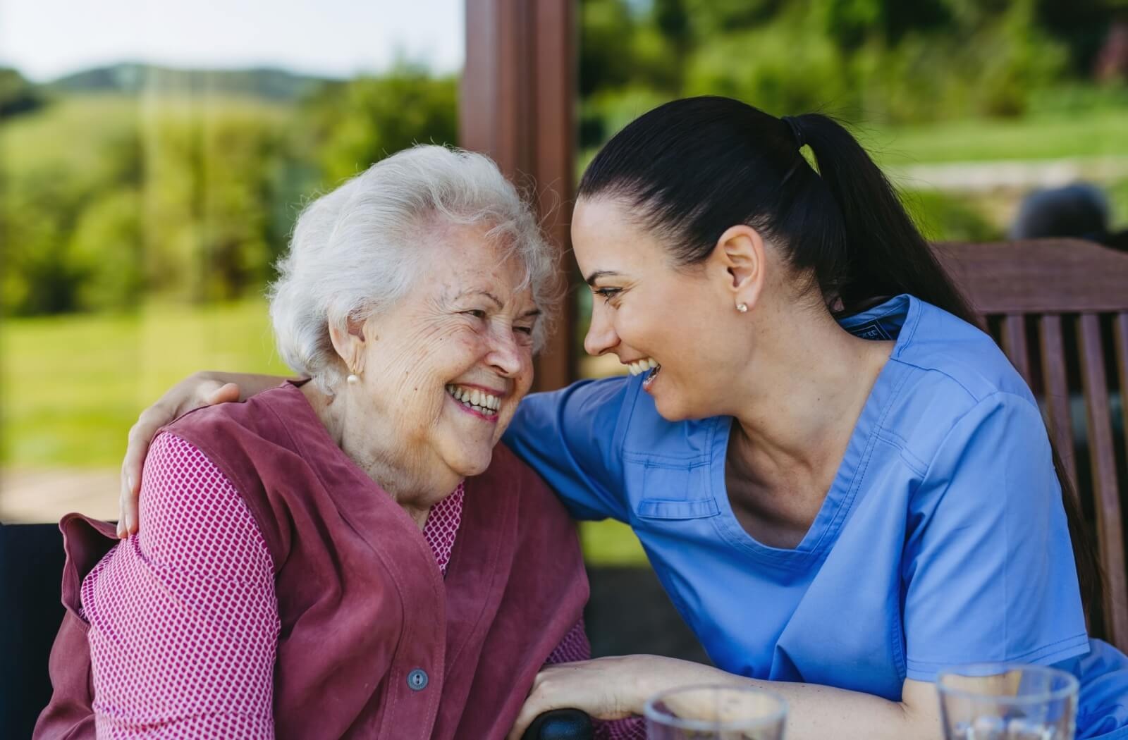 A caregiver and older adult laugh while sitting in a sunroom in memory care