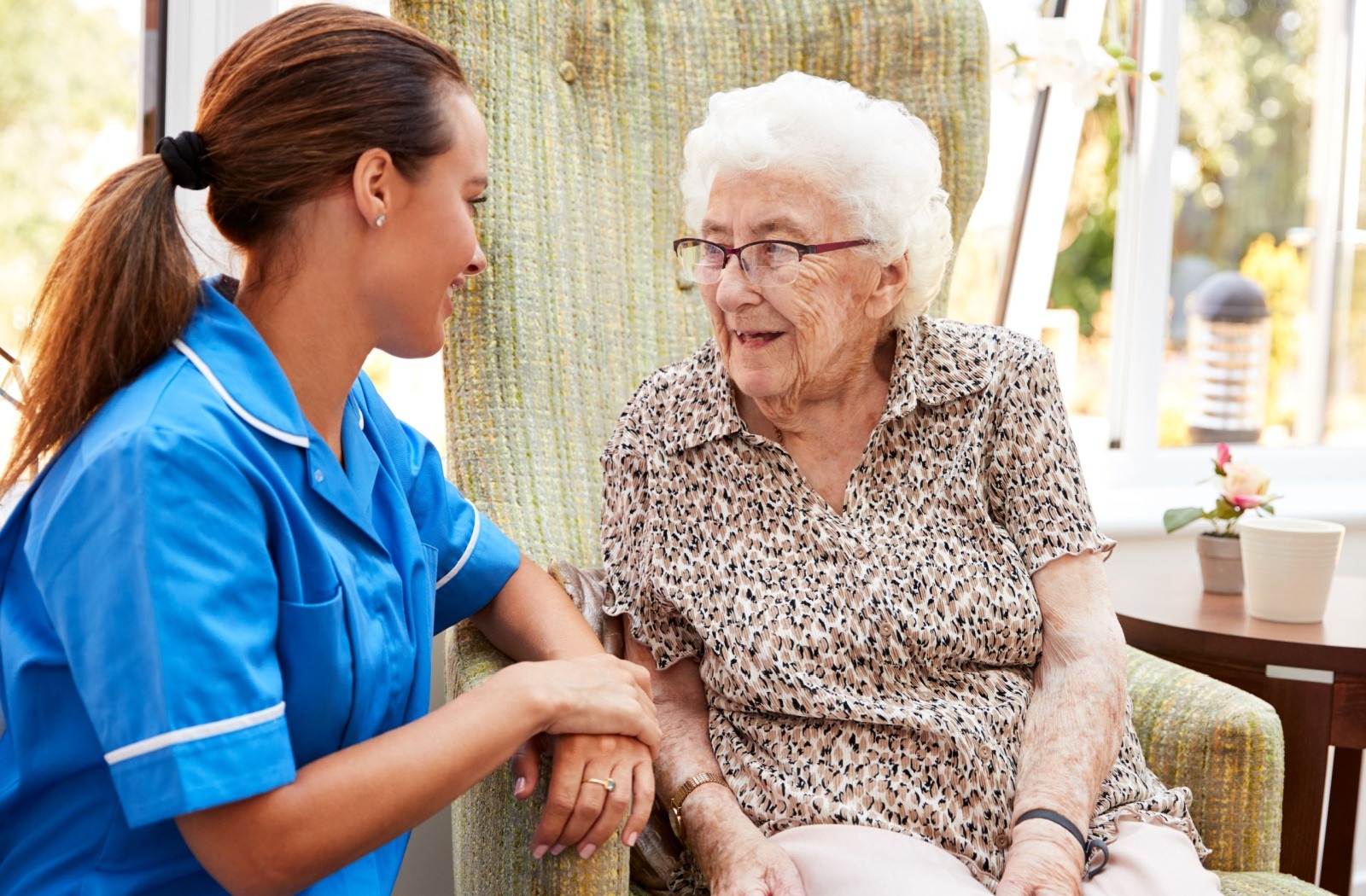 A caregiver smiles and talks to a memory care resident sitting in an armchair.
