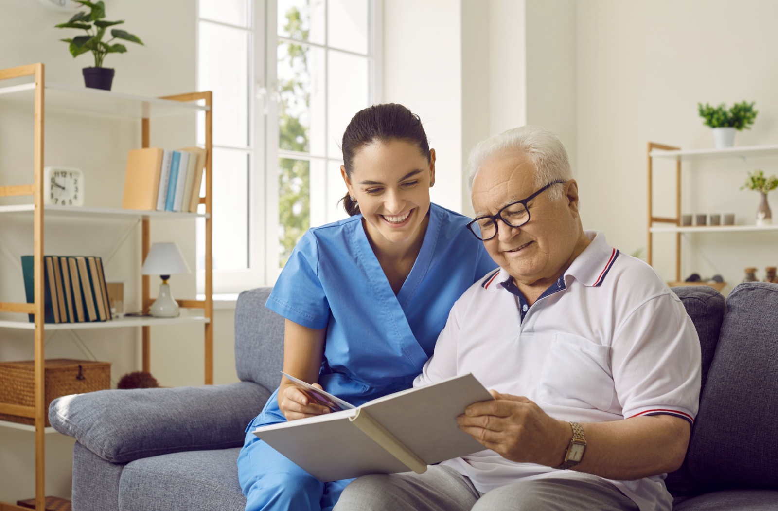 A memory care caregiver sits on a couch with a resident with dementia, looking through a photo album.
